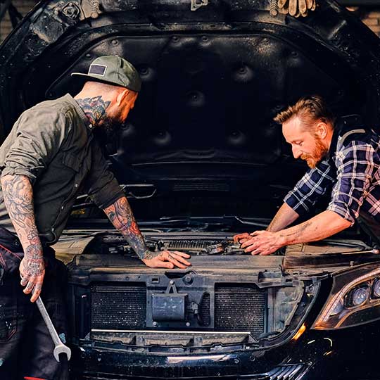 Das Bild zeigt zwei Männer, die an einem Auto arbeiten. Die Motorhaube des Autos ist geöffnet und die Männer scheinen den Motorraum zu inspizieren oder zu reparieren.