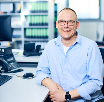 Markus Pelz sitzt lächelnd am Schreibtisch in einem Büro trägt ein hellblaues Hemd und eine Brille
